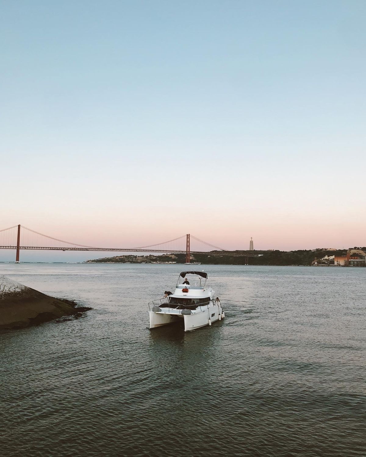 A SkipperPedro catamaran on the Tagus at dusk, with the 25 de Abril bridge behind